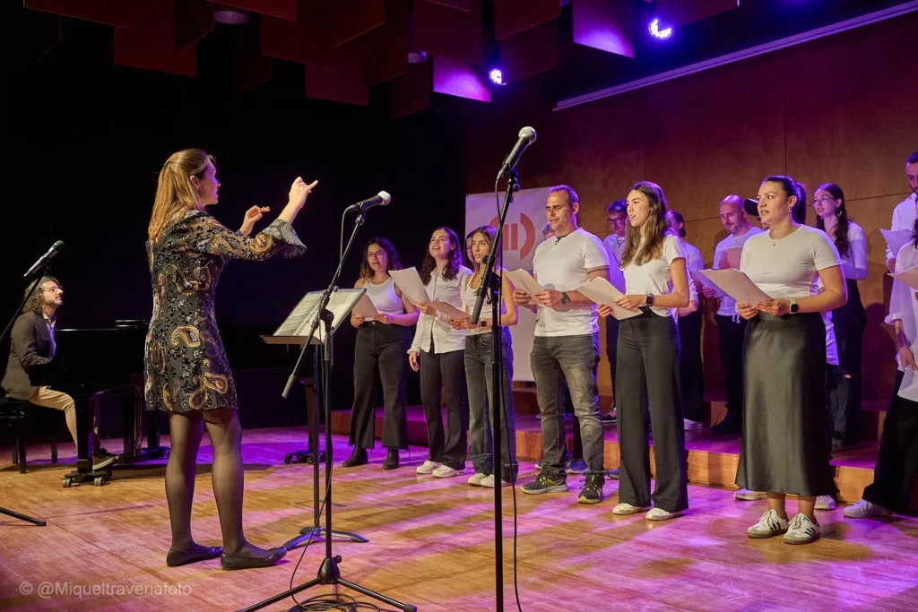 Lana Kovačević dirigeix el cor damunt l'escenari, amb els cantaires amb les partitures a les mans i un pianista al fons.