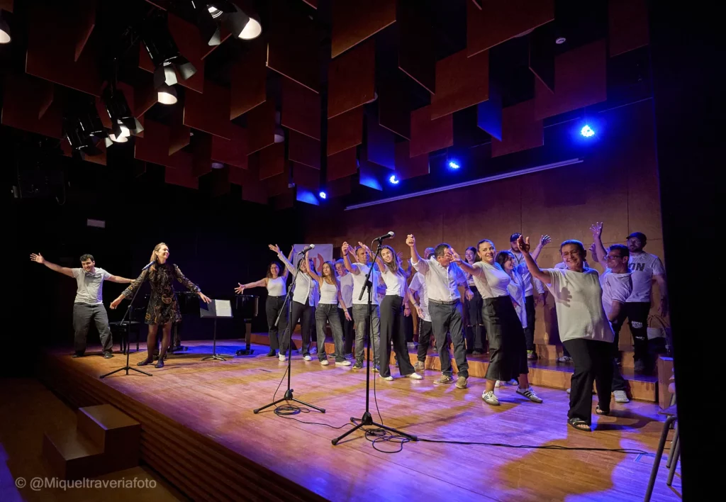 El cor al complet, amb els braços oberts i les mans agafades, saluda al final del concert damunt l'escenari.
