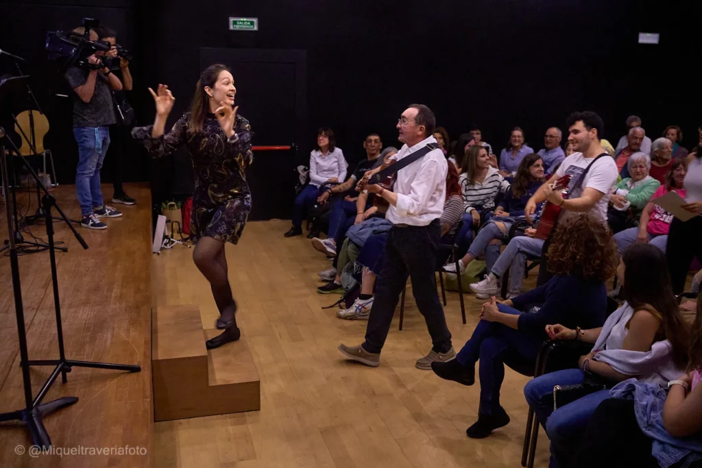 Lana Kovačević dirigeix des d'una tarima mentre un guitarrista toca caminant entre les cadires del públic.