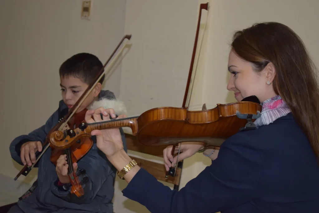 Lana Kovačević toca el violí al costat d'un infant que la imita amb el seu instrument, tots dos amb l'arc a l'aire.