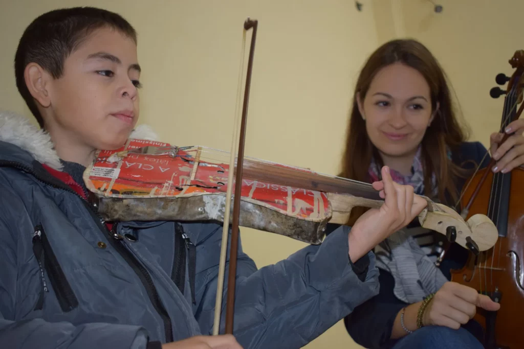 Un noi toca un violí construït amb una llauna reciclada mentre Lana Kovačević l'observa somrient, amb un violí de fusta a les mans.