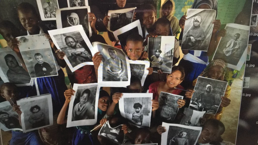 Un grup d'infants somrient sostenen en alt retrats fotogràfics en blanc i negre, en una aula de Gàmbia.