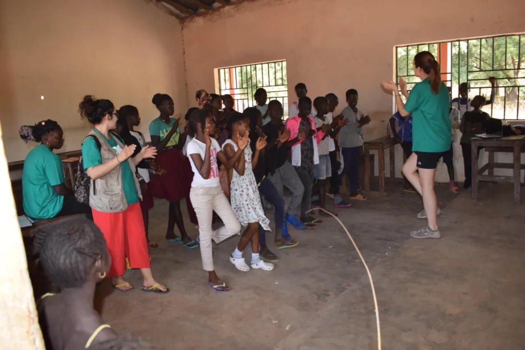 Un grup d'infants i de voluntàries piquen de mans dretes dins d'una aula, seguint una monitora.
